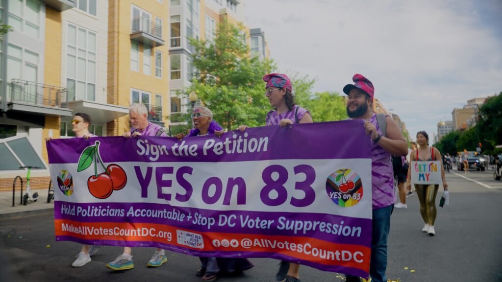 Five people holding a banner while walking in a pride parade. The banner reads: "Sign the petition. Yes on 83. Hold politicians accountable + stop DC Voter Suppression"