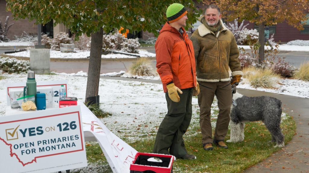 Two people and a dog standing beside a sidewalk next to a sign that reads "Yes on 126 OPEN PRIMARIES FOR MONTANA"