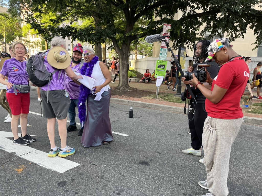 People vs. Politics crew (right) in Washington, D.C., films Initiative 83’s Lisa Rice at the 2024 Pride Parade, with audio and camera gear in use and pedestrians in the background.