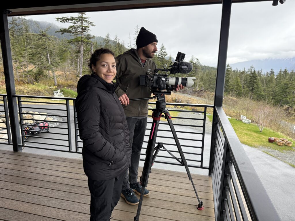 Two crew members from People vs. Politics film in Alaska on a cloudy day, capturing early efforts of the “No on Two” campaign to preserve Alaska's Final Four voting, with mountains and deep green pine trees in the background.