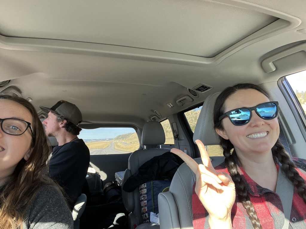 People vs. Politics associate producer Mary Claire smiles from the passenger seat during a Montana road trip with 2 other crew members. They smile as we can see the open road and plains through the rear window.